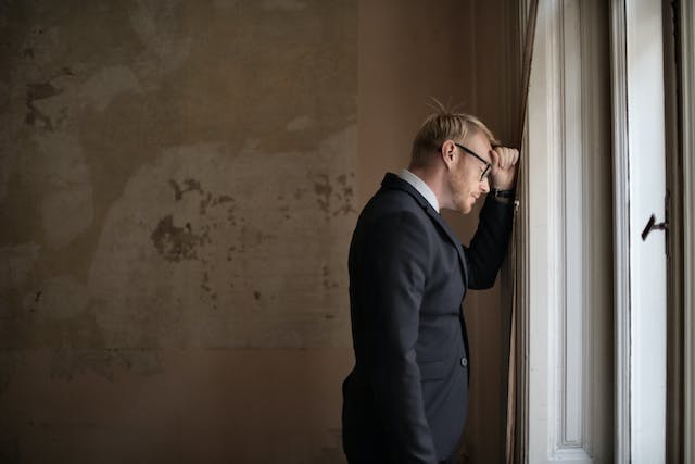 A forlorn businessman in a room, leaning on a window sill